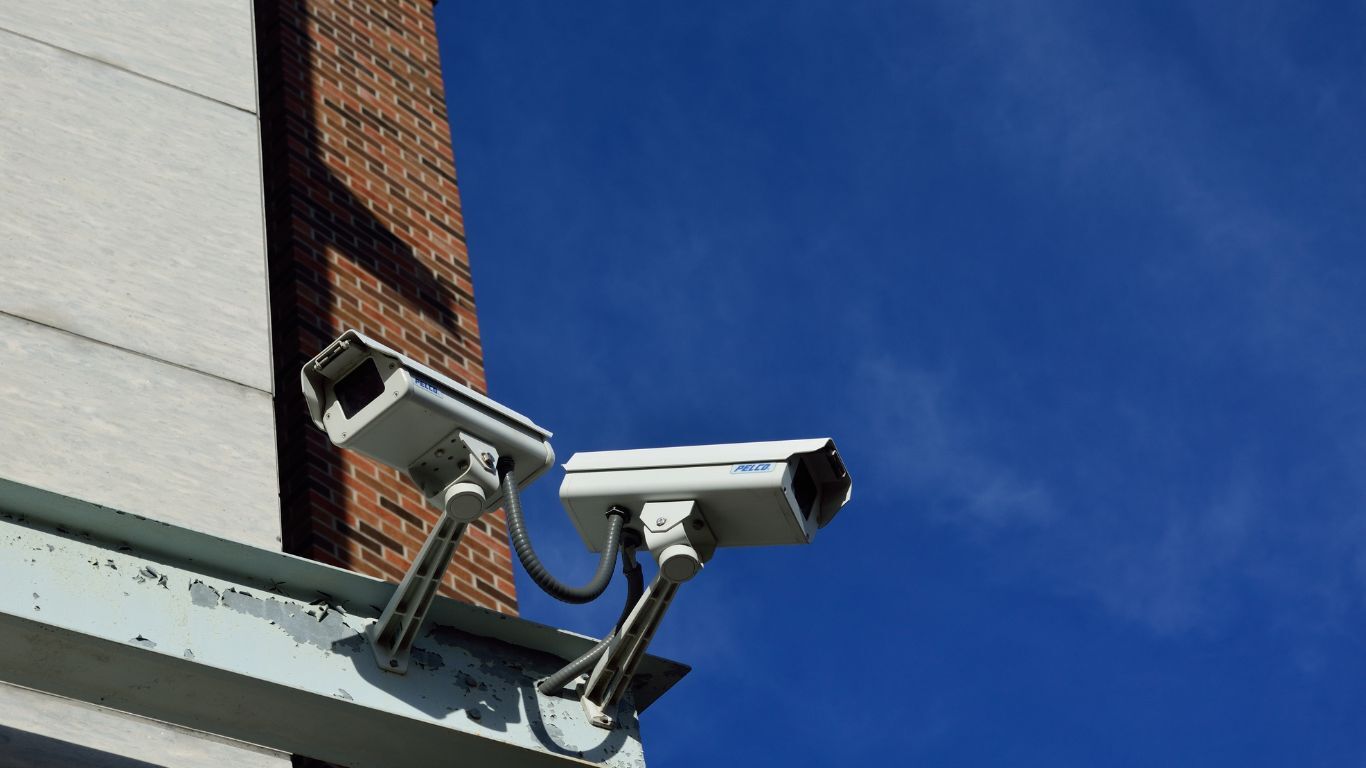 Two security cameras on the side of a building. There is a brick facade and blue sky in the background.