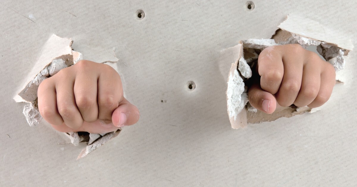 Two fists punching through a broken drywall wall, exposing the screw pattern on the studs behind — illustrating how the Denver break-in crew entered through a shared wall instead of a door.